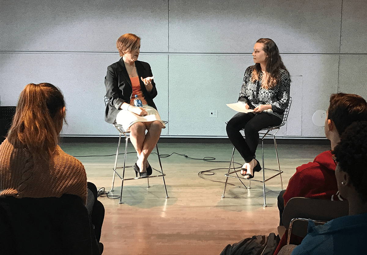 Rutter speaks with a student, both seated on stools in front of a room.