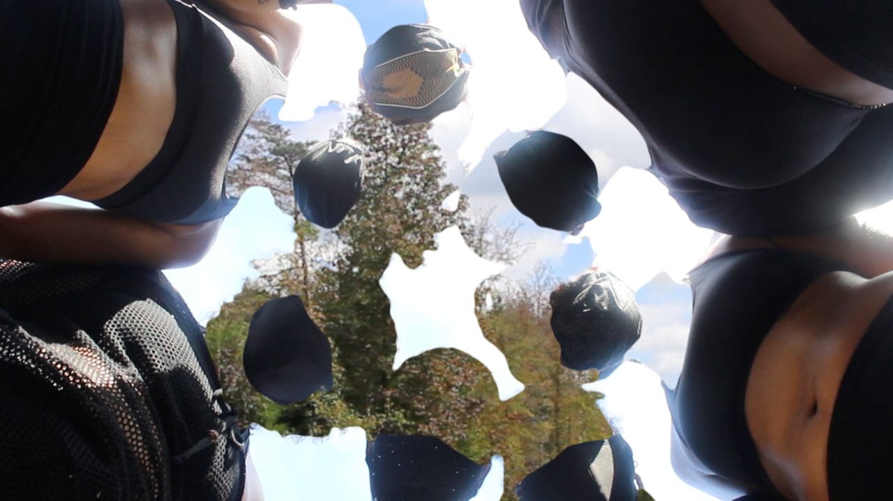 A photo of 7 figures in a circle looking down into the camera. They are Black people and are dressed in black to various degrees with black masks. Their faces and necks are replaced with an image of a tree line.