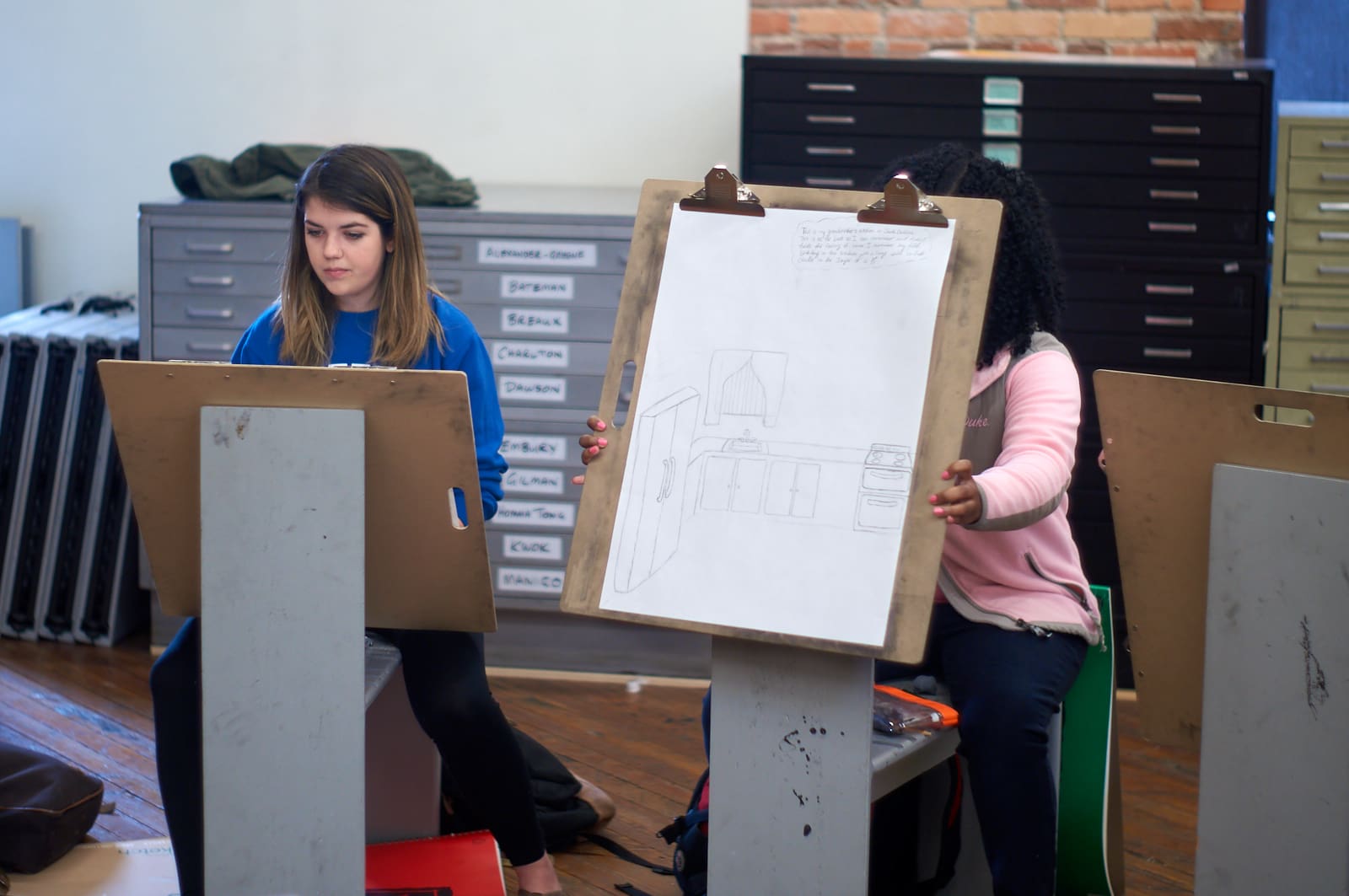 A student holds her drawing in front of her as she describes the room she drew