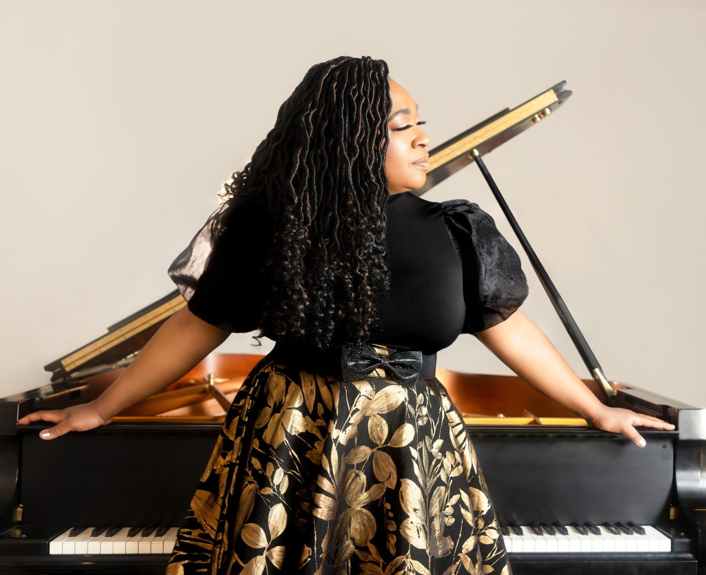 A women with long dark curly hear facing to the side as she leans against a piano