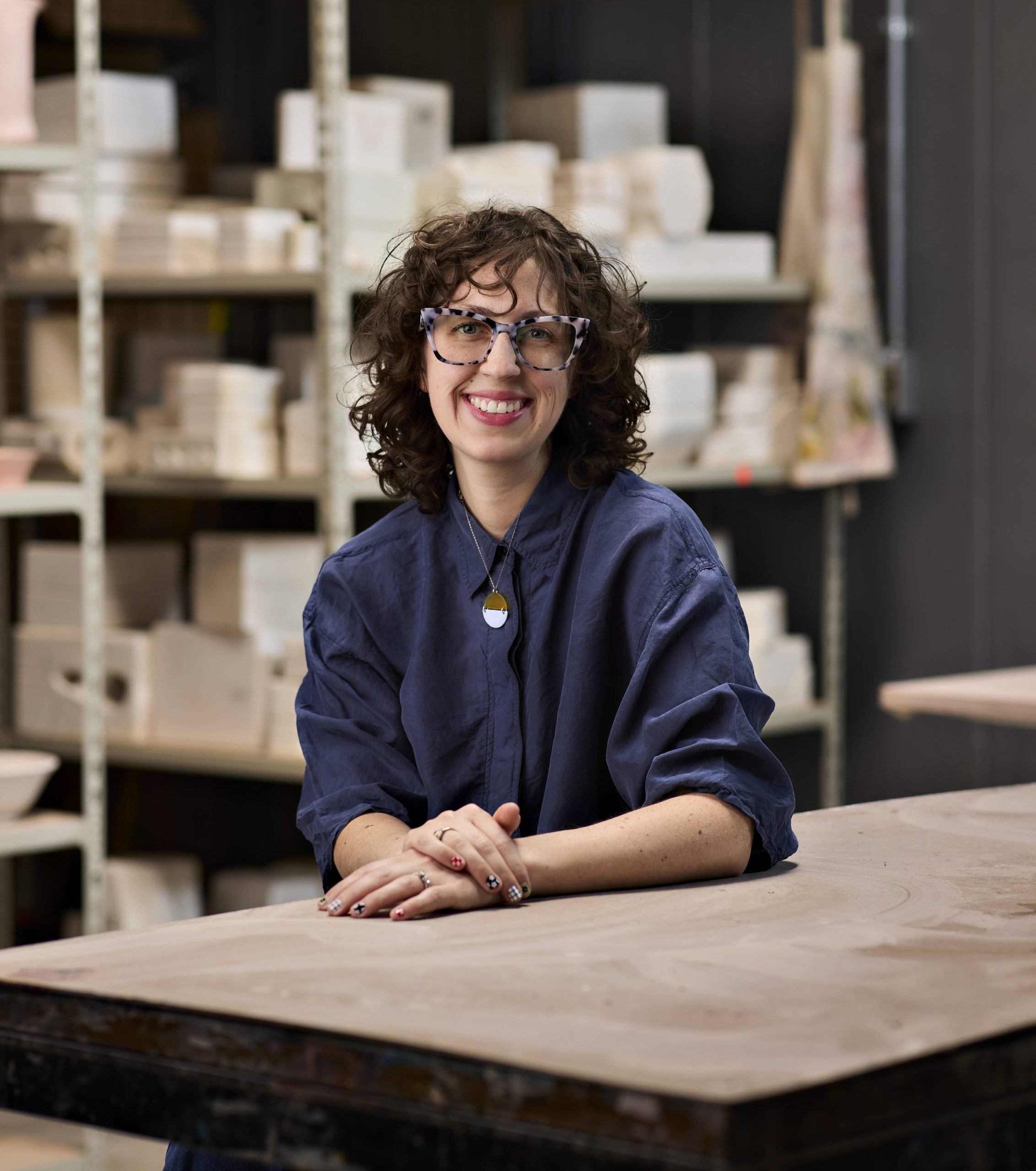 Woman with curly brown hair sitting at ceramics table with ceramics on a shelf behind her