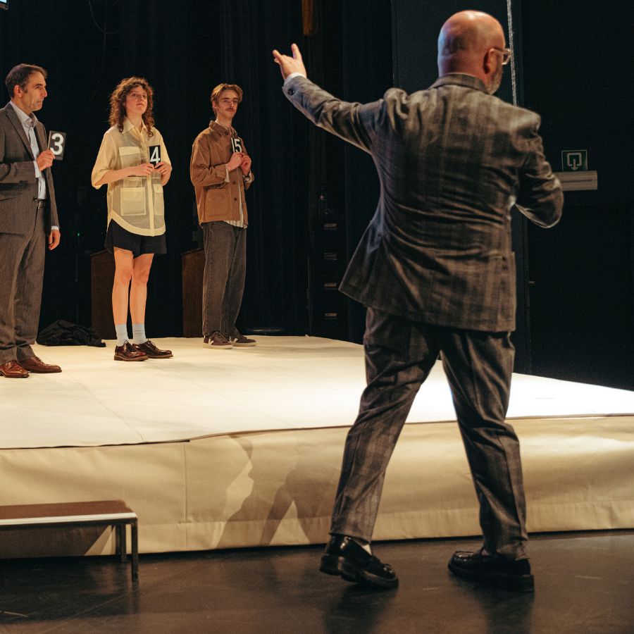 Man pointing at 3 people upon a stage. He is wearing a brown suit