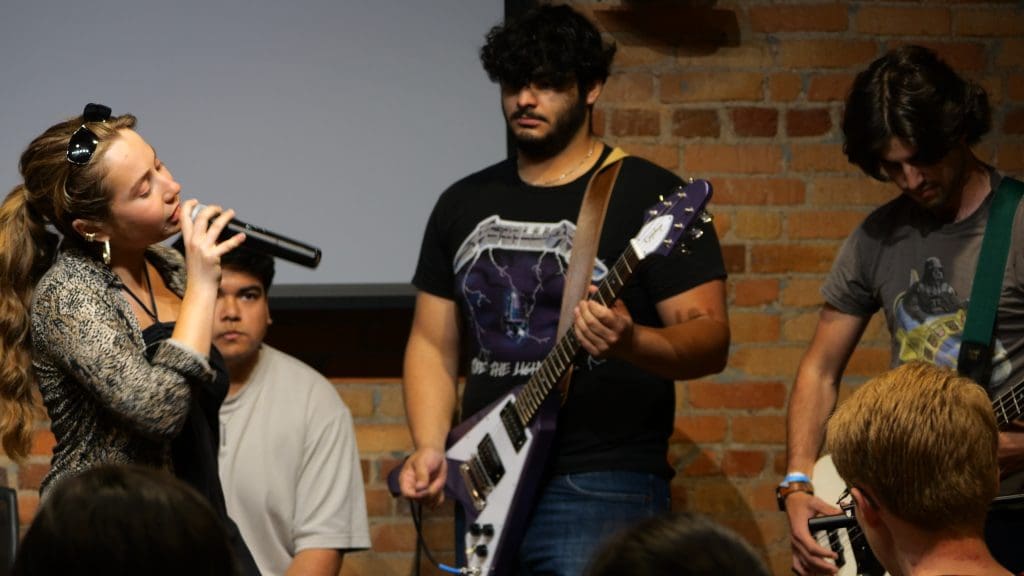 Three students performing. One young adult women holding a mic, two young adult men playing the bass and guitar