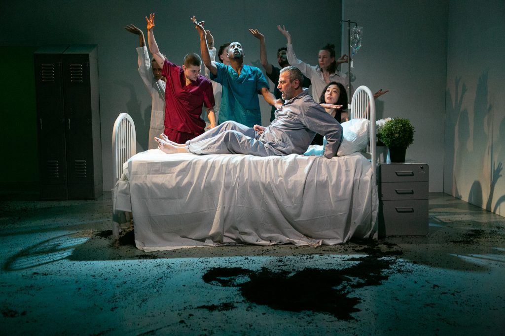 Several medical professionals stand in scrubs of different colors and lift one hand to the sky behind a man in a hospital bed