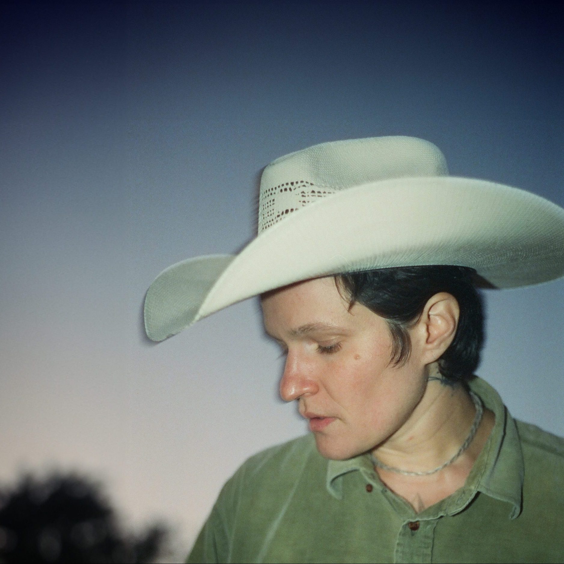 Person with short hair wearing green shirt and cowboy hat against a melancholy sunset.