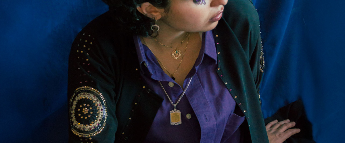 A person dressed in a purple button up shirt underneath a beaded dark emerald long sleeve jacket paired with blue denim jeans