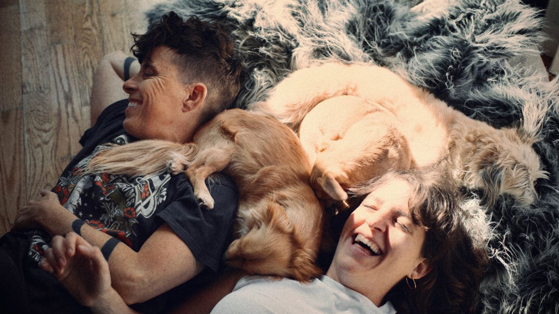 Two people are lying on a fluffy rug, laughing joyfully. Two small dogs snuggle with them, creating a warm, cozy, and affectionate scene.