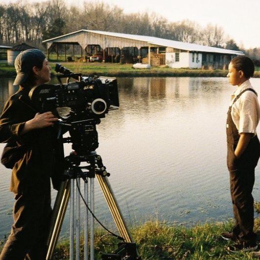 A young adult student working a film camera pointed at a young child next to a pond
