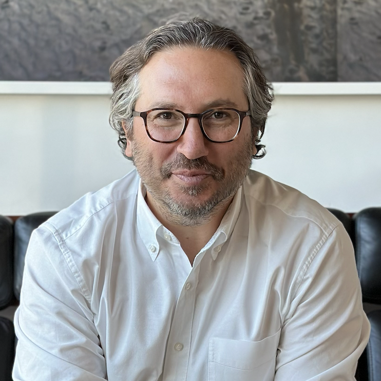 A white man with white hair wearing glasses and a white button up and jeans sitting on a couch